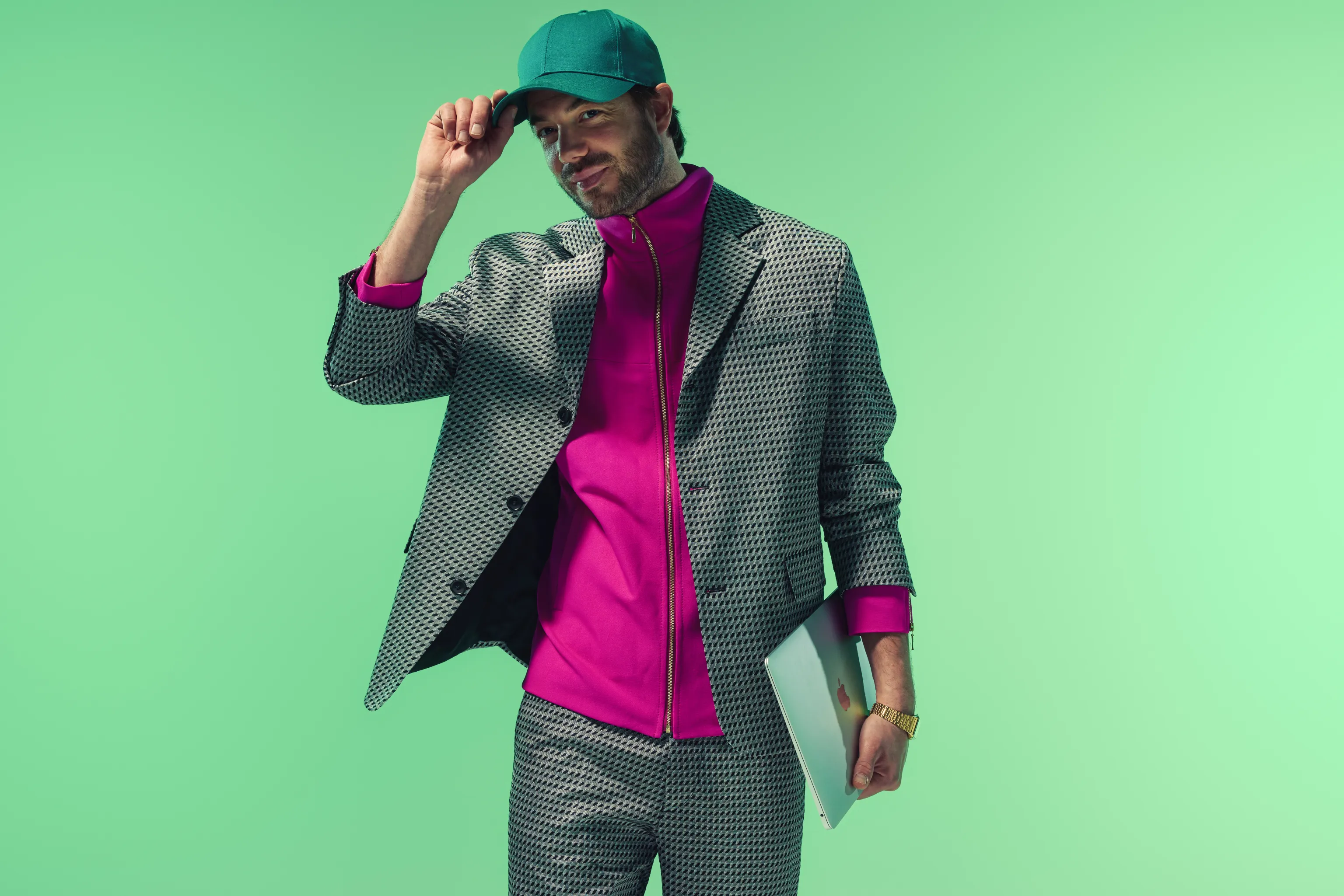 Een man in een modern grijs pak, een felroze jas en een groene pet, die een laptop vasthoudt en lachend naar de camera kijkt, als symbool voor een mensgerichte benadering van gedragsverandering.