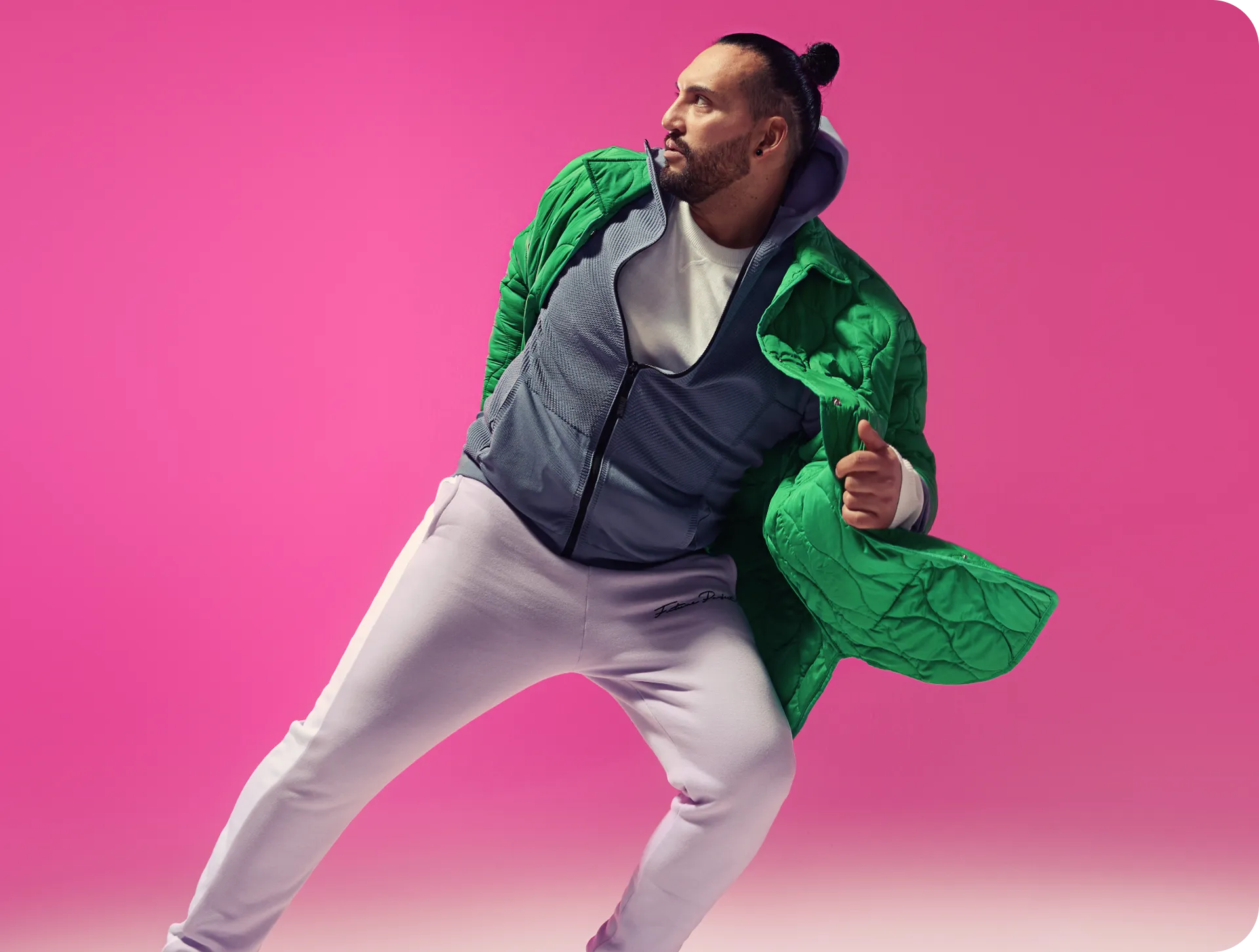Een stijlvolle man in een dynamische pose, gekleed in een groene gewatteerde jas en lichte sportkleding tegen een felroze achtergrond, wat een voorbereiding op ethische disruptie uitbeeldt.