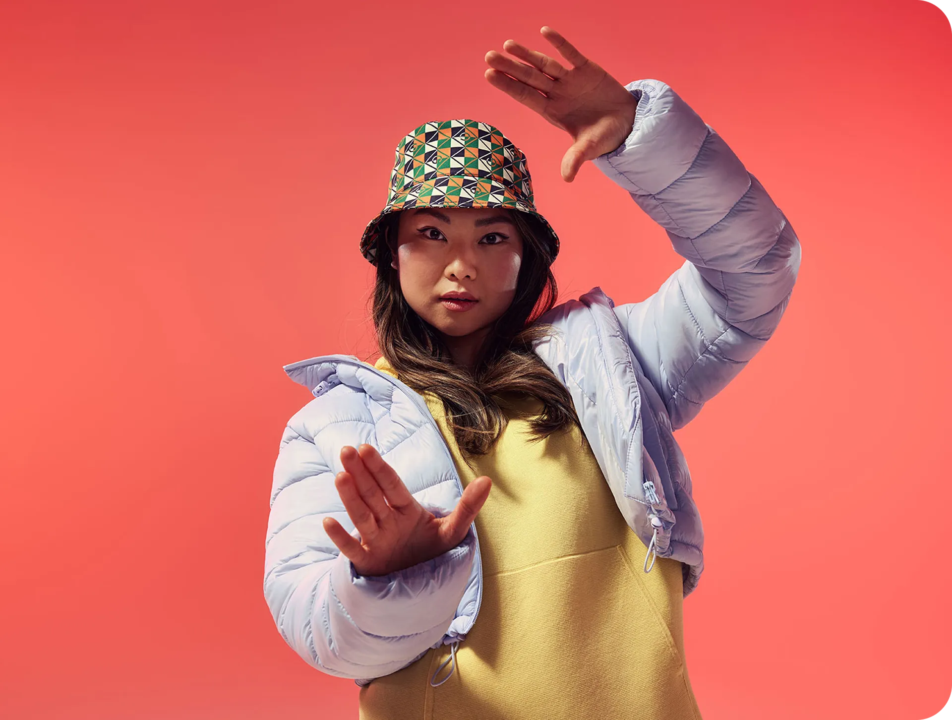 Een vrouw in een lichtblauwe pufferjack en een bucket hat met patroon, die haar handen omhoog houdt alsof ze wil begeleiden of ondersteunen, wat een toegewijde programmamanager uitbeeldt.