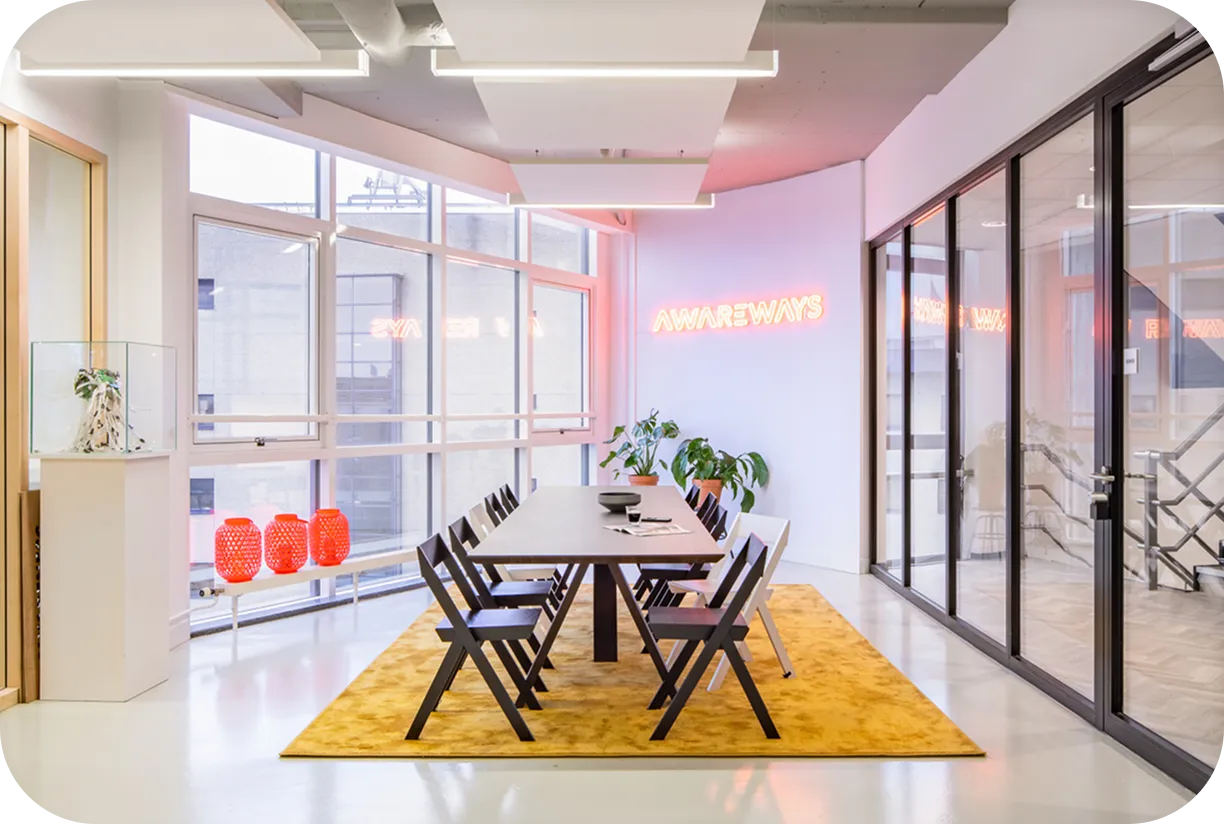 A modern office meeting room with a long wooden table, black chairs, and a yellow rug. Large windows let in natural light, and a neon sign on the wall reads “AWAREWAYS.” Three red lamps sit on a bench near the window, and a green plant adds a touch of color.