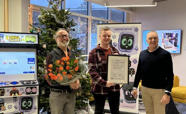 Foto van Maarten Timmerman, Jeroen Koppe en een vertegenwoordiger die zojuist het ISO 9001-certificaat en bloemen heeft overhandigd aan Awareways. Ze staan voor een kerstboom en een arcadekast in het kantoor van Awareways.