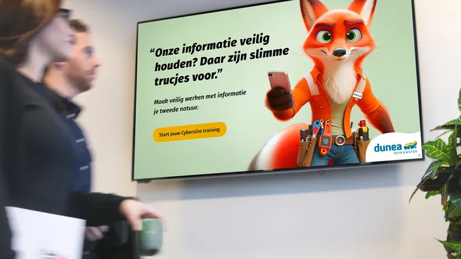 Een digitaal scherm in een kantoor toont een cartoonvos met een smartphone en een boodschap over informatiebeveiliging en het starten van een cybersecuritytraining. Op de voorgrond lopen medewerkers langs het scherm.